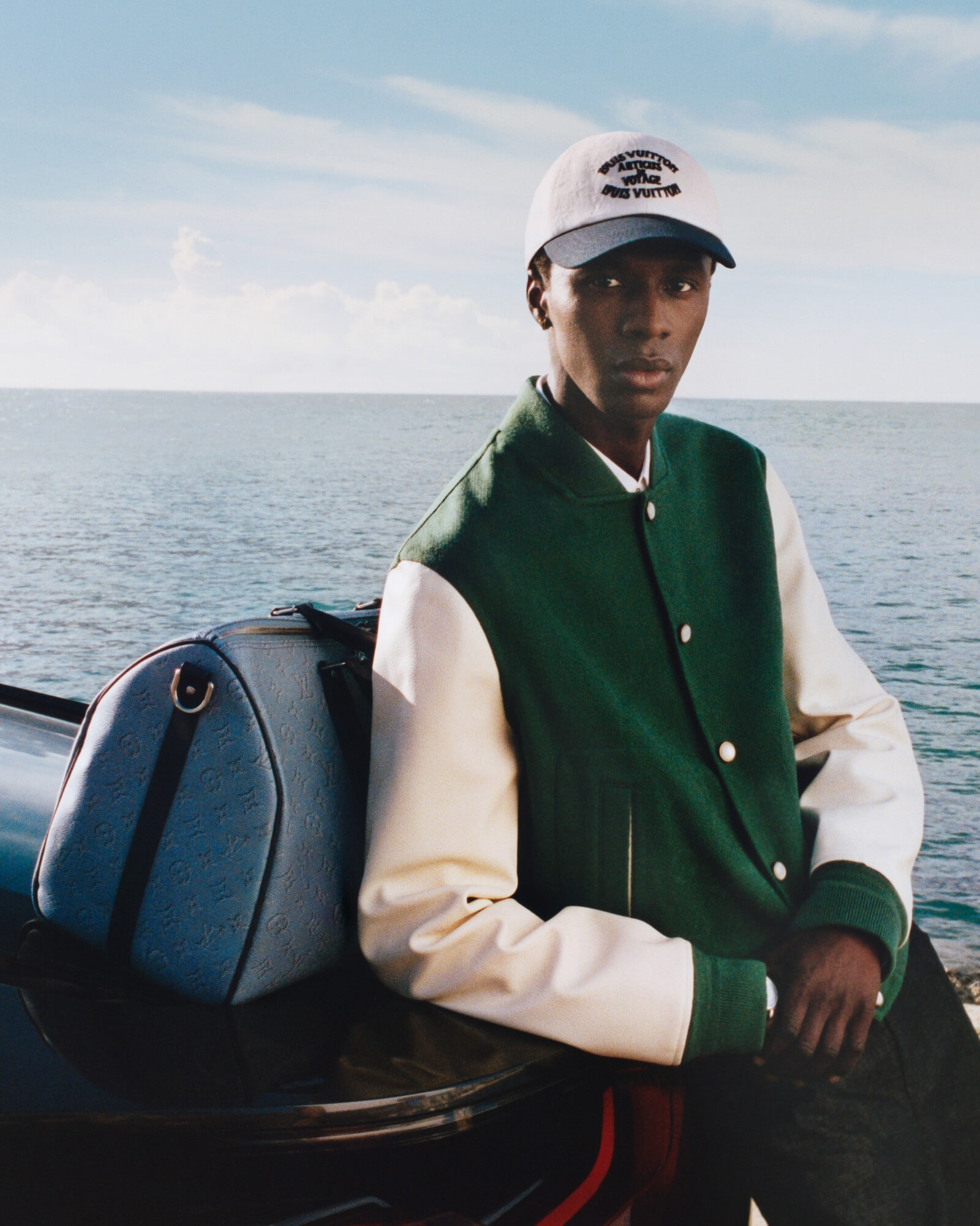 Model wearing a Louis Vuitton cap with a varsity jacket beside a monogram travel bag.
